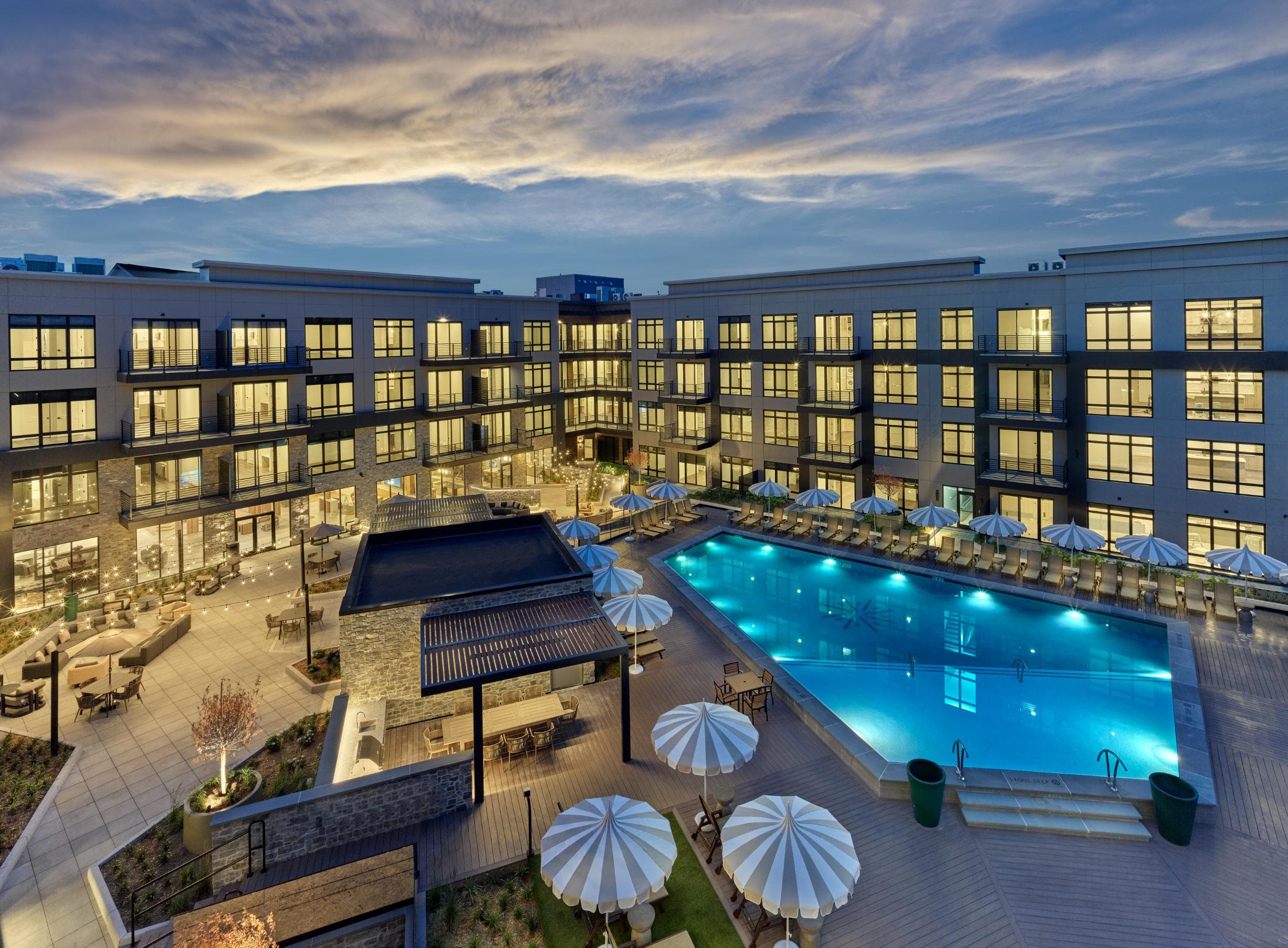 A large swimming pool surrounded by lounge chairs and umbrellas is the centerpiece of a resort-style courtyard in a luxury apartment rental community