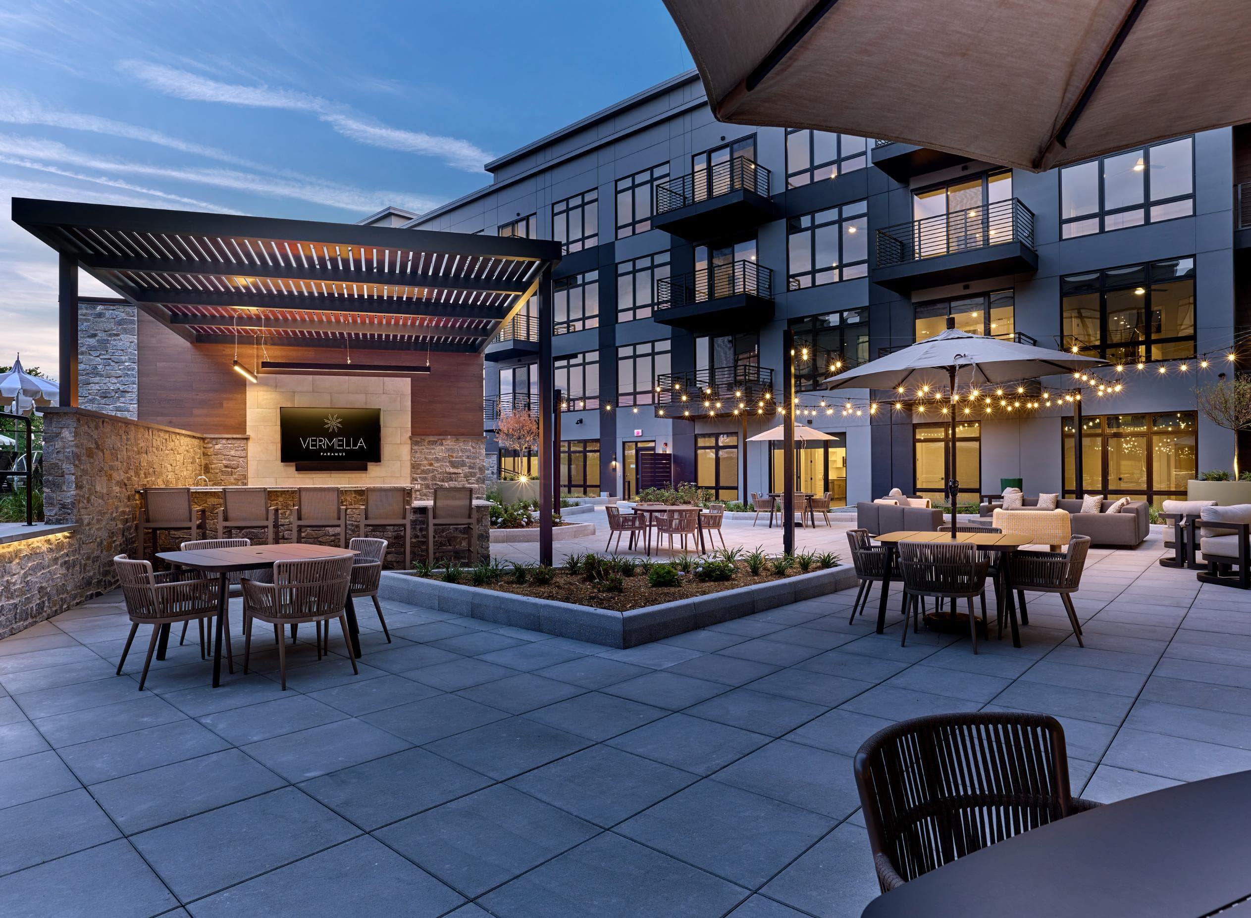 A modern outdoor dining area with tables and chairs is illuminated by string lights.