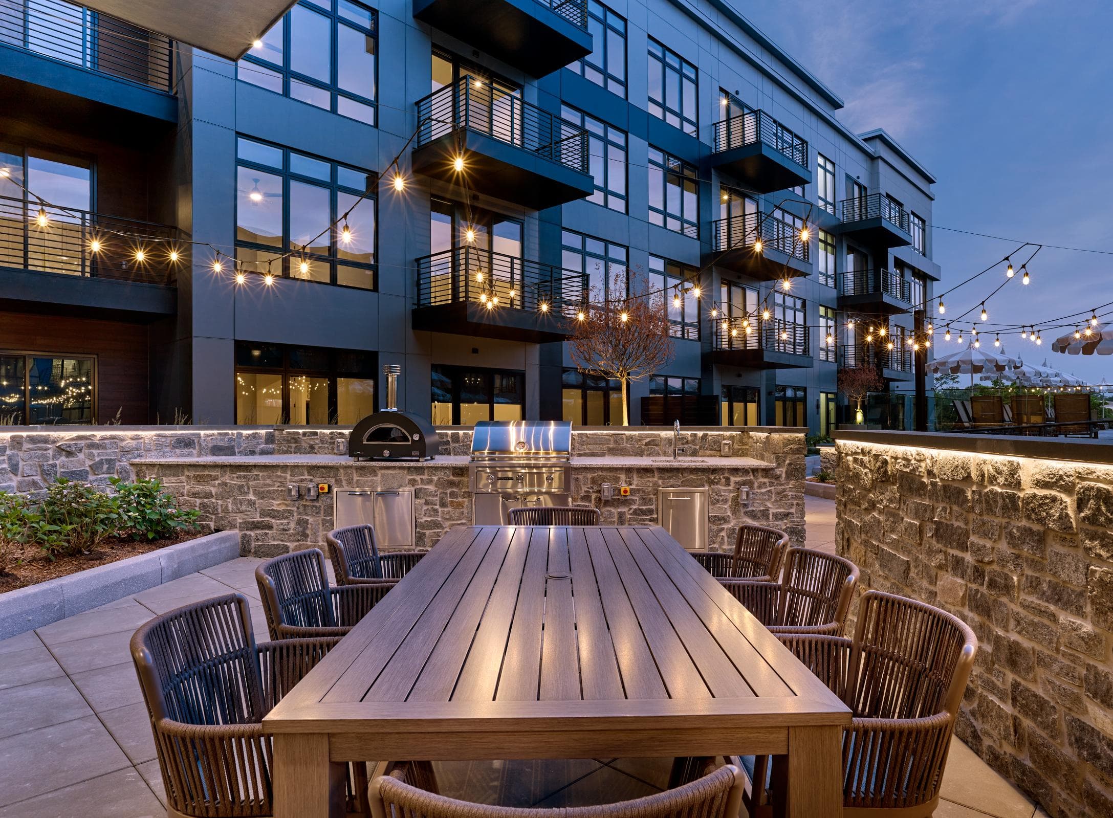 A wooden table surrounded by chairs in front of a building with lights strung up.